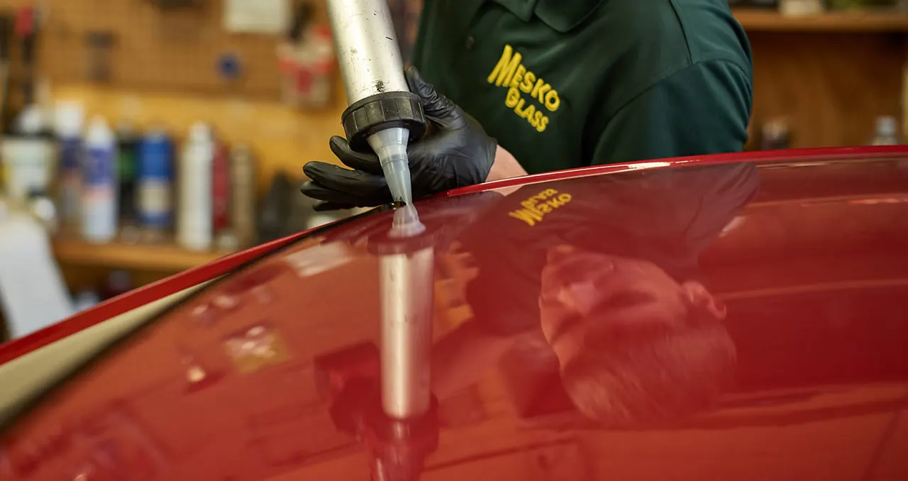 Gloved hands running a bead of urethane along the windshield opening of a red car in the glass shop