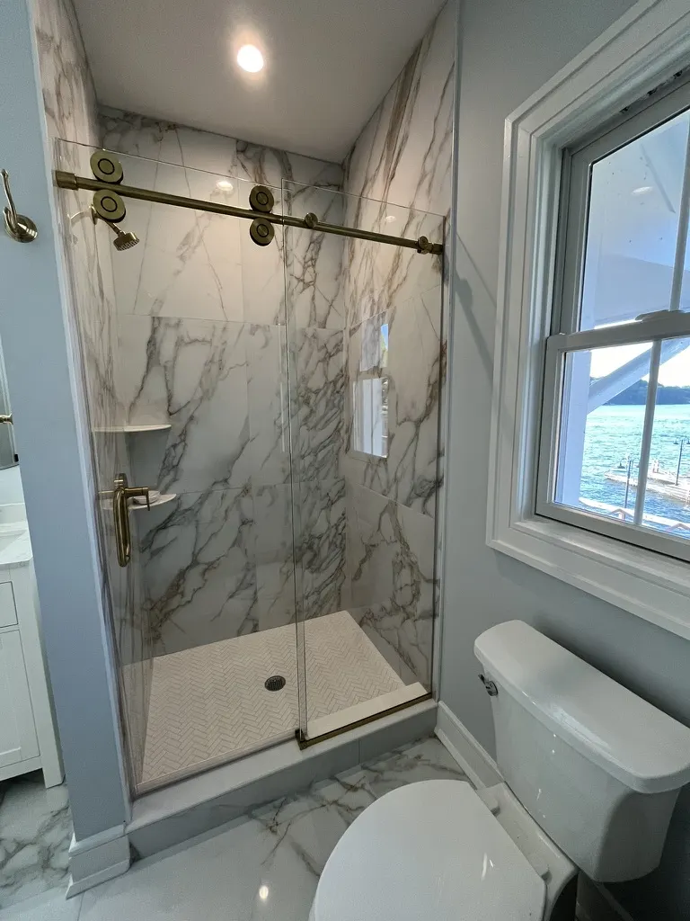 Sliding shower door on antique-brass barn hardware in a white marble-look bathroom with a lake view through the window.
