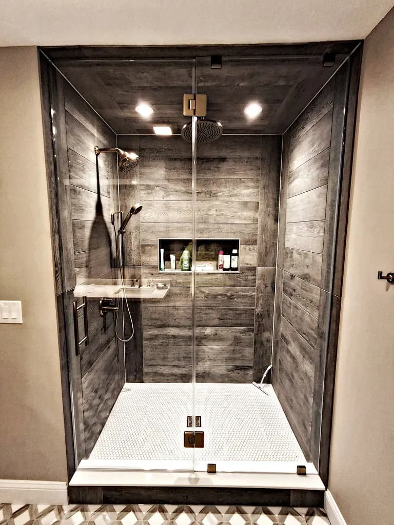 Frameless glass shower enclosure against gray barnwood-look plank tile, with a penny-round floor.